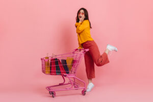 Surprised girl in pink culottes posing with trolley full of multi-colored packages with new clothes.